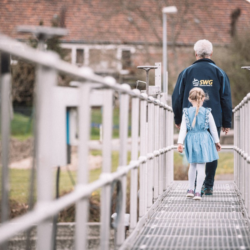 Ein Kind läuft hinter einem Mitarbeiter der Stadtwerke bei einer Führung durch die Kläranlage über eine Brücke.