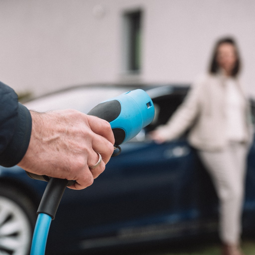 Eine Hand steckt einen Ladeanschluss in ein Elektroauto, im Hintergrund lehnt eine Frau an dem Fahrzeug.