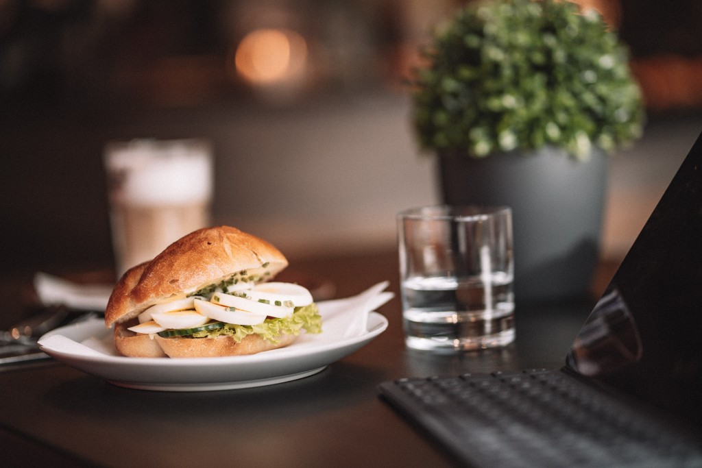 Brötchen mit Salat und Ei belegt, ein Glas Wasser, ein Capuccino und eine kleine Grünpflanze auf einem schwarzen Tisch